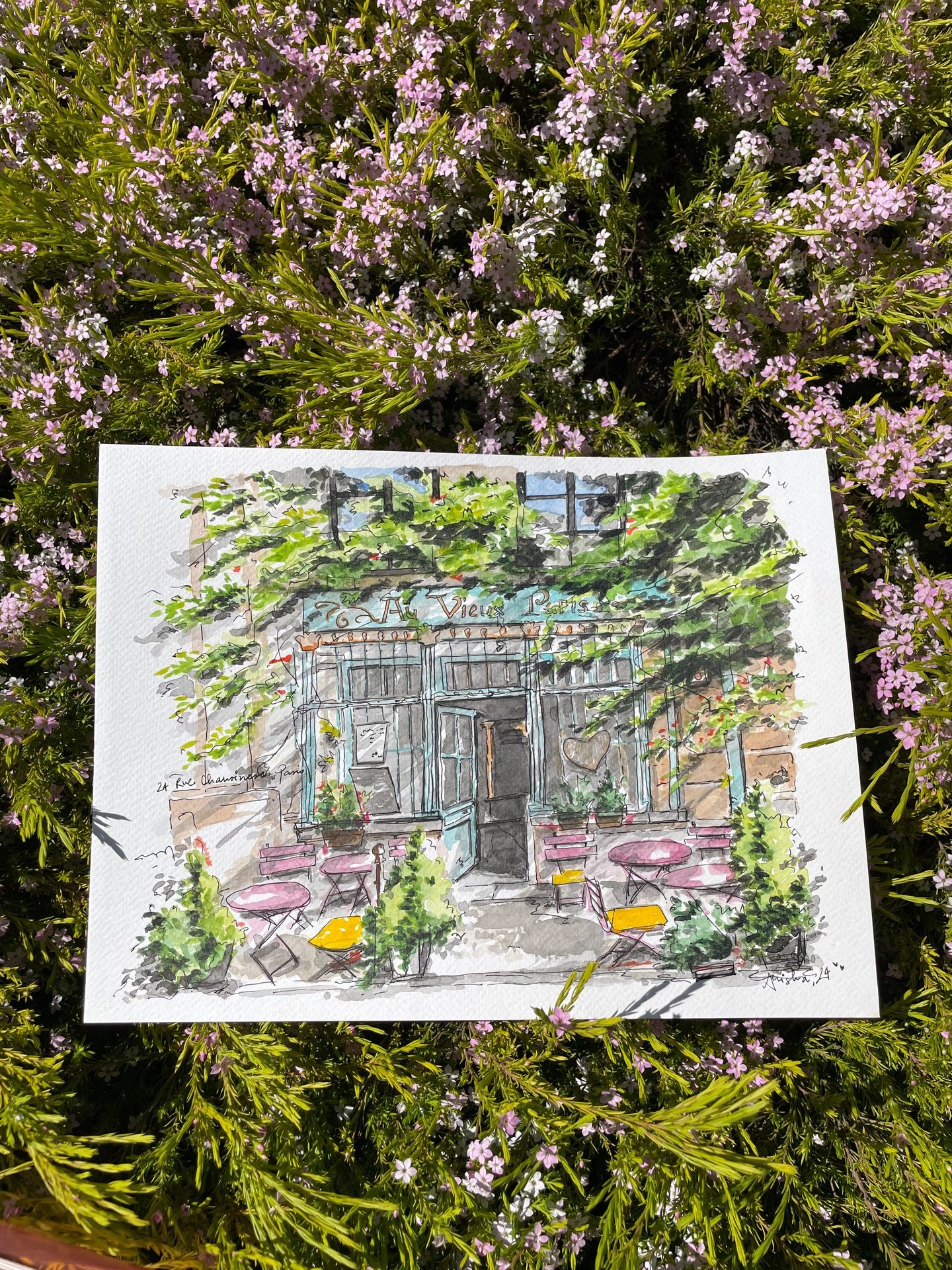 Watercolor painting of a café with outdoor seating, placed on blooming pink and green foliage.