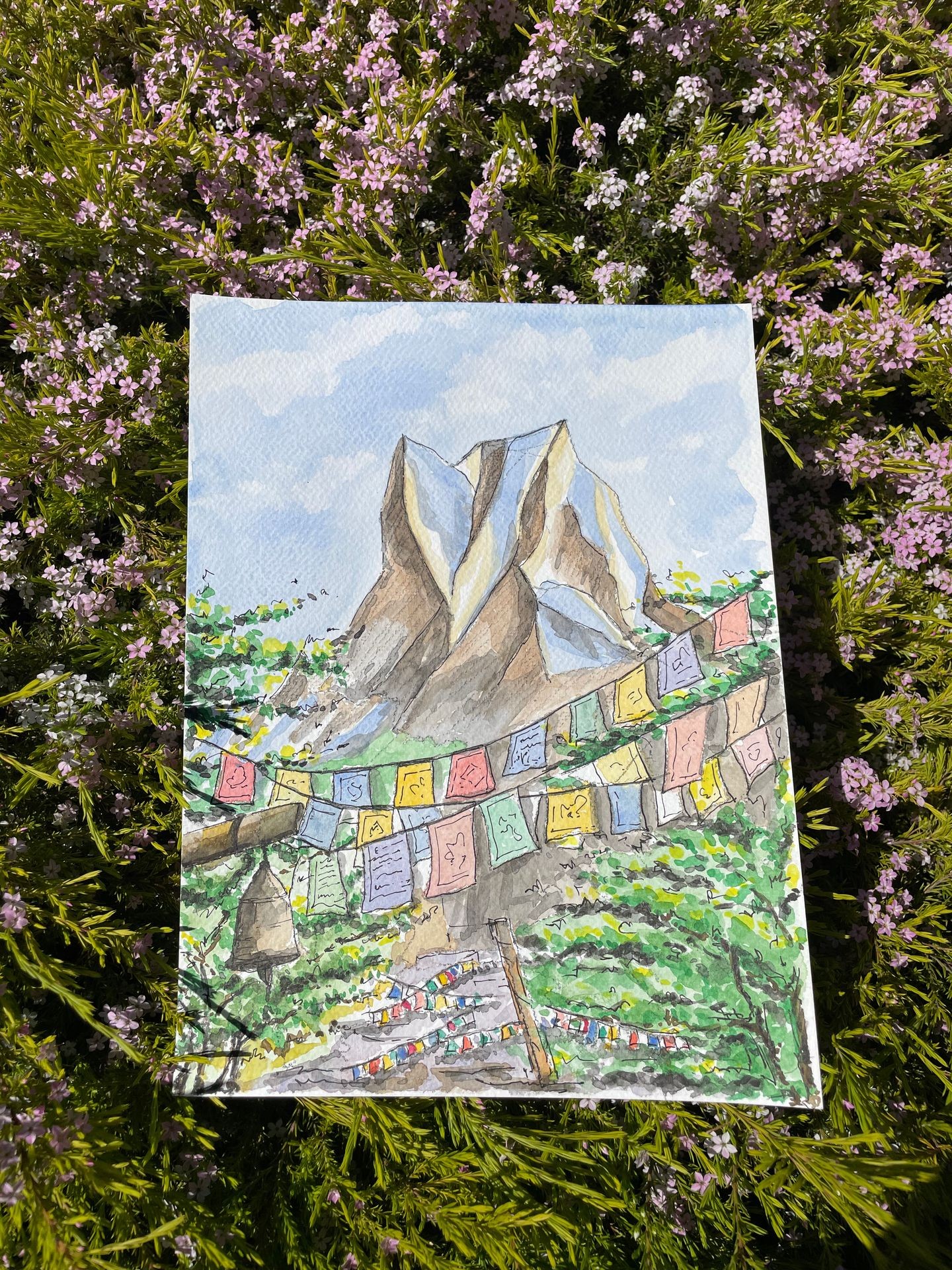 Painting of a mountain with colorful prayer flags in a grassy field with small pink flowers.