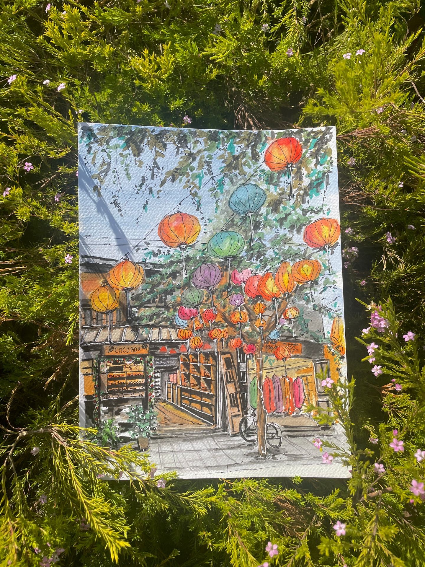 Colorful painting of a street scene with lanterns, displayed outdoors in green bushes.